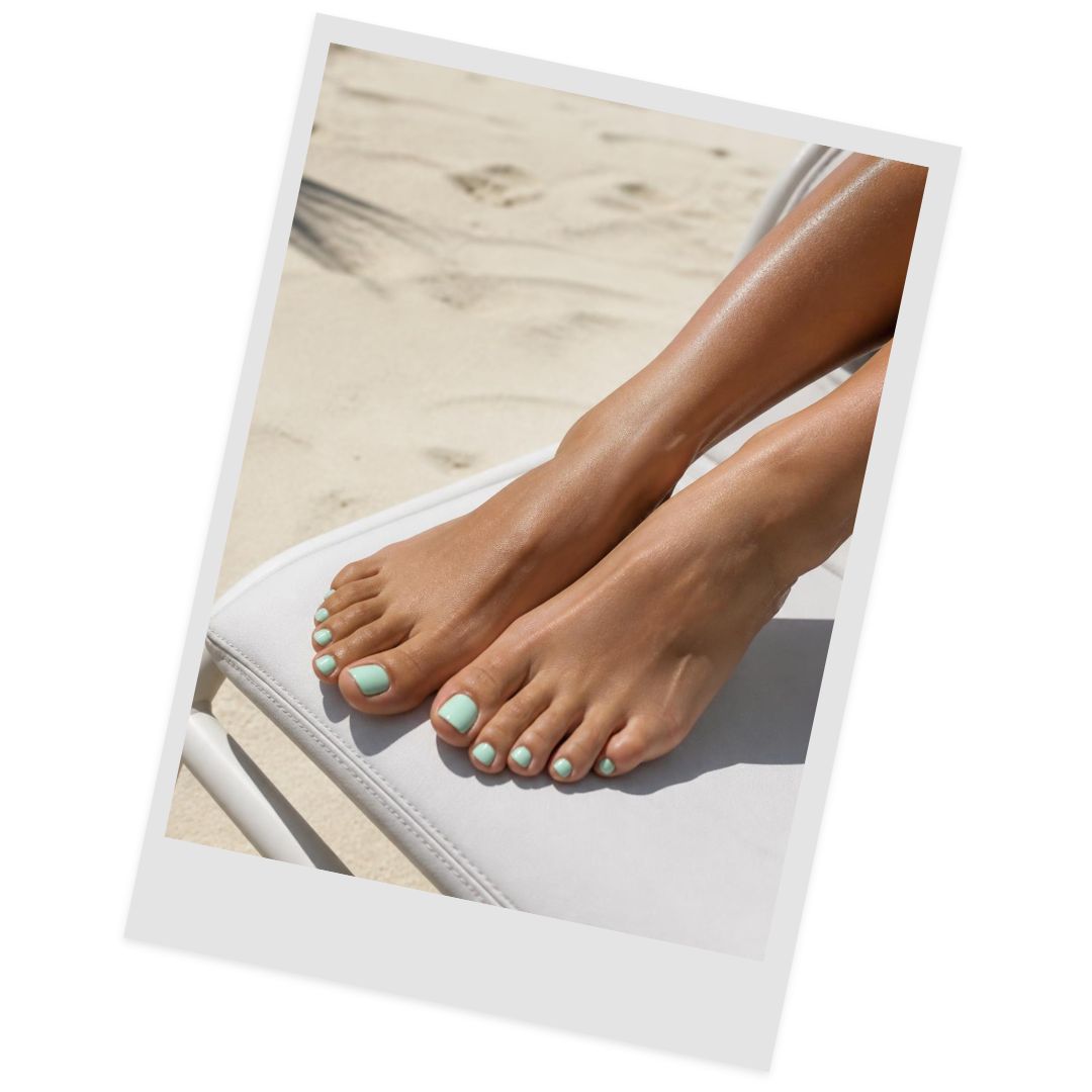 Feet with mint green toenail polish resting on a white lounge chair at the sandy beach.