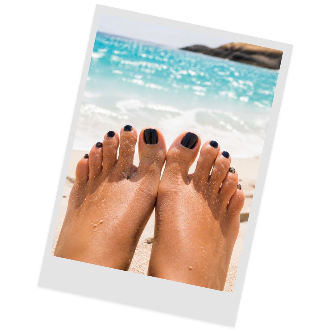 Feet with black toenail polish resting on sandy beach, turquoise ocean in the background.